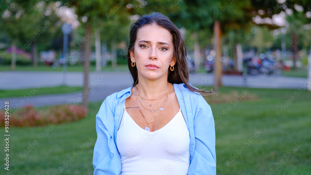 A young, beautiful businesswoman with brown hair is looking at the camera with a sad expression in a tree-filled park on a sunny summer day A young, beautiful businesswoman with brown hair is looking at the camera with a sad expression in a tree-filled park on a sunny summer day