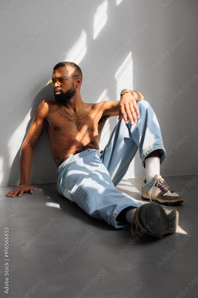 Shirtless black man in sunglasses sitting in sunlight near wall Shirtless black man in sunglasses sitting in sunlight near wall