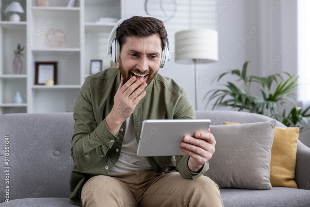 Man sitting on sofa wearing headphones while watching a funny video on tablet. He laughs and touches his face, engrossed in content. Relaxed home setting with modern decor.