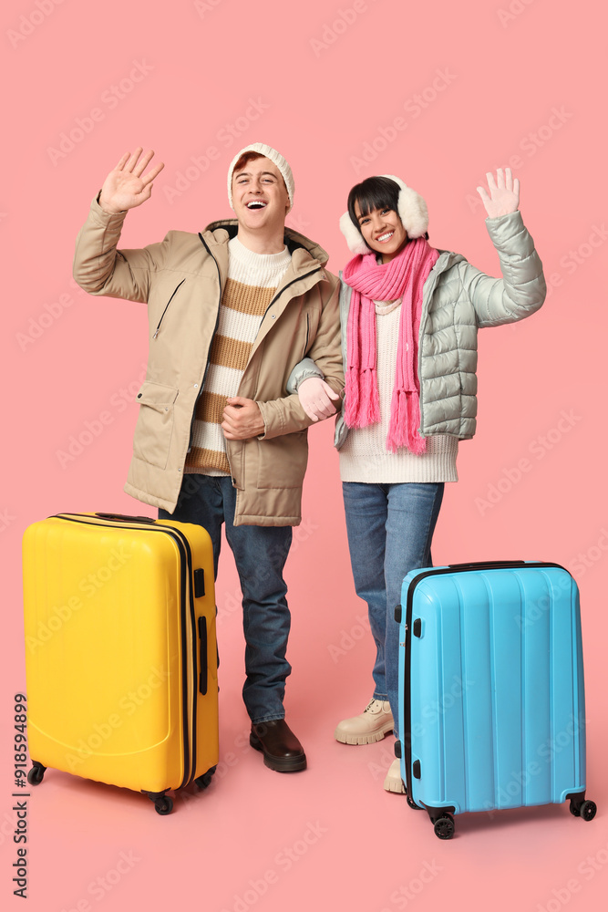 Beautiful young happy couple with suitcases waving hands on pink background. Winter vacation concept Beautiful young happy couple with suitcases waving hands on pink background. Winter vacation concept