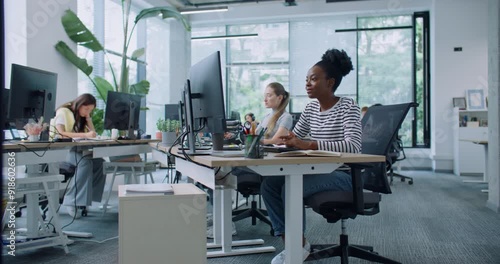 Team of mixed-raced workers sitting at desks and using corporate computers in modern white office. Camera zooming in on beautiful woman typing text on keyboard. Enjoying working in financial company.