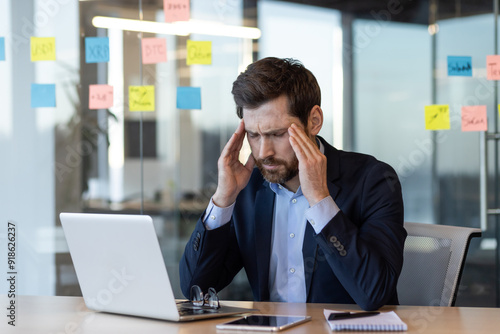 Stressed businessman sitting at office desk working on laptop, holding his head, experiencing a headache or frustration. Office environment with sticky notes and documents. Concept of work stress and