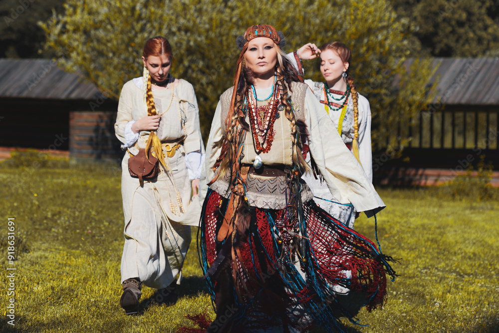 Naklejka premium Young girls and a shaman woman in folk costumes in a meadow, in a village among wooden architecture, Slavic or Nordic style