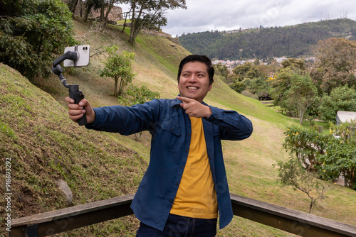 Smiling Latino man dressed casually, vlogging outdoors in the city of Cuenca with his smartphone and stabilizer, talking to the camera