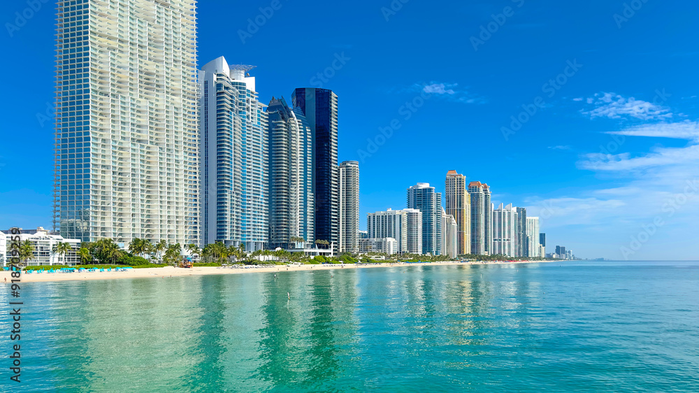 Naklejka premium Miami Sunny Isles Beach. Skyscrapers on bright sunny Florida summer day. Ocean with blue clear sky. Travel location for vacation with luxury architecture. City with sand beach and clear blue water.