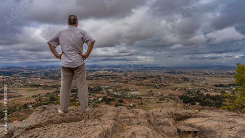 A man stands on the edge of a cliff, looking at the panorama of the valley in front of him. Rural houses among the trees, fields, red-soil dirt roads are visible. Clouds, sky. Mountains on the horizon photo