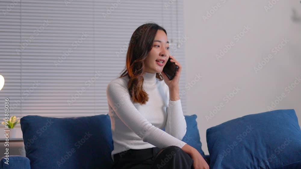 Young professional woman sits on a sofa, talking on her phone. She receives surprising news, checks her phone in disbelief, and crosses her arms Young professional woman sits on a sofa, talking on her phone. She receives surprising news, checks her phone in disbelief, and crosses her arms