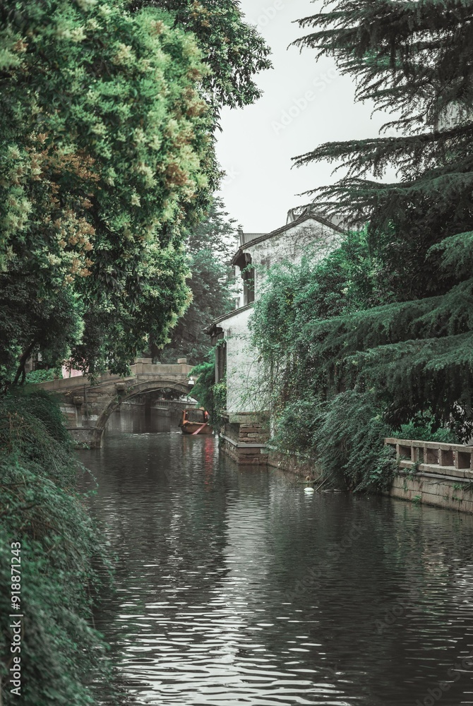 A canal in Suzhou, China, with a stone bridge and a rowboat. Lush greenery lines the banks and a white-walled building peeks through the foliage.