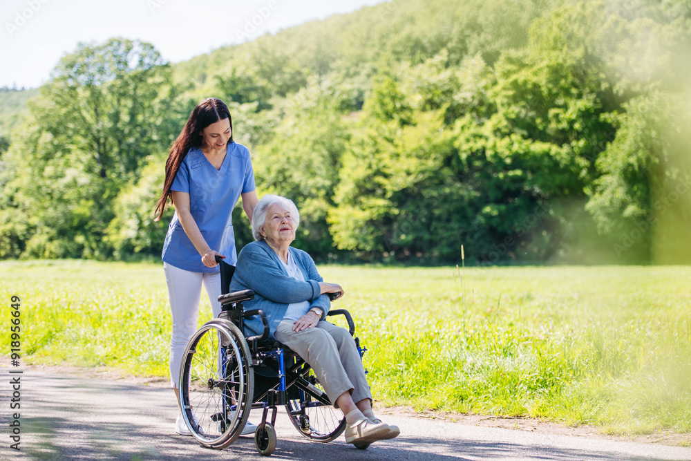 Female caregiver pushing senior woman in wheelchair. Nurse and elderly woman enjoying a warm day in nursing home, public park. Female caregiver pushing senior woman in wheelchair. Nurse and elderly woman enjoying a warm day in nursing home, public park.