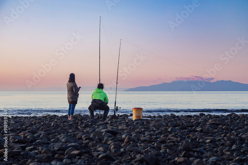 People fishing along the shoreline of the Firth of Thames at sunset at Orere Point, Auckland, New Zealand. In the background is the Coromandel Peninsula.