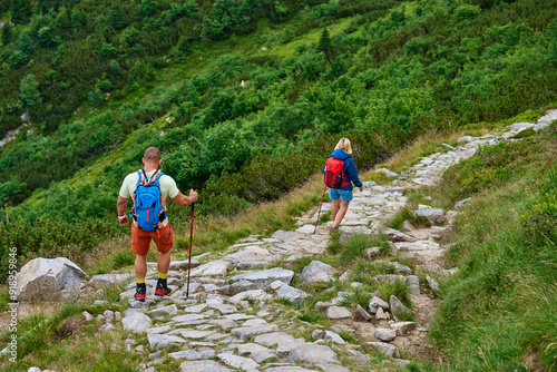 Back view of two hikers with backpacks and trekking poles descending rocky mountain trail. Mountain hiking concept. Active recreation in nature