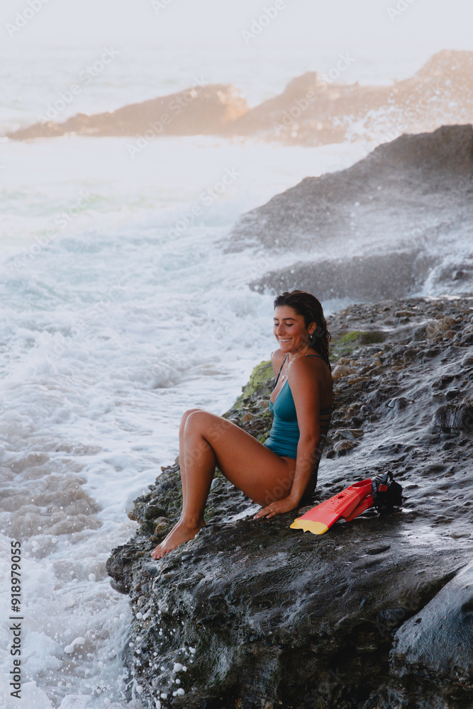 Woman sitting with swim gear on cliff next to waves breaking Woman sitting with swim gear on cliff next to waves breaking