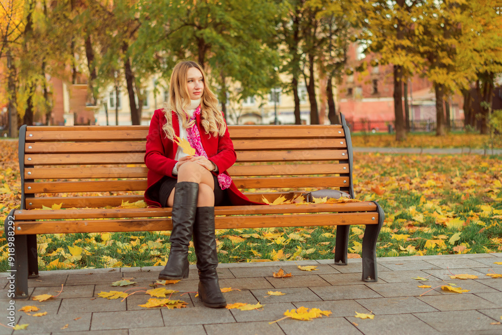 A girl in a red coat sits on a bench in an autumn park. She holds yellow leaves in her hand. A girl in a red coat sits on a bench in an autumn park. She holds yellow leaves in her hand.