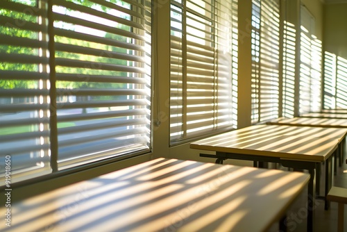 Sunlight streaming through window blinds in empty classroom, casting patterned shadows on desks. Bright and serene atmosphere highlighting calm and peaceful learning environment