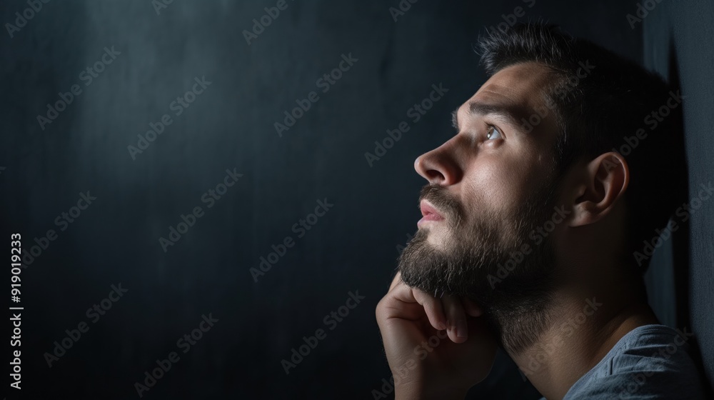 A man with a beard rests his chin on his hand, gazing thoughtfully upward in a softly lit, dark environment, deep in contemplation about his thoughts A man with a beard rests his chin on his hand, gazing thoughtfully upward in a softly lit, dark environment, deep in contemplation about his thoughts