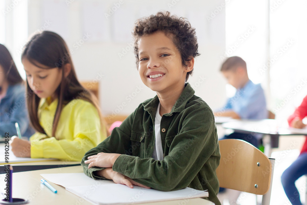 Smiling schoolboy sitting at desk in classroom and smiling, classmates writing in copybooks on the background. Education, learning and people concept Smiling schoolboy sitting at desk in classroom and smiling, classmates writing in copybooks on the background. Education, learning and people concept