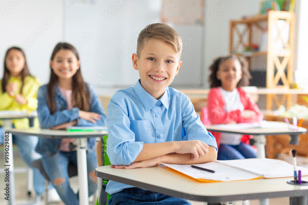 Fototapeta premium Happy European schoolboy sitting at school desk and smiling at camera with diverse classmates on background. Smart kids and education concept