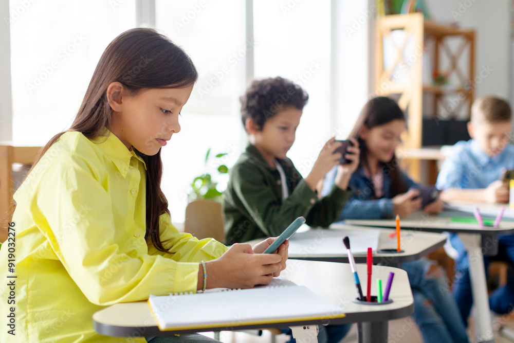 Multiethnic kids pupils having break and using their smartphones, sitting at desks in classroom, playing games, focus on girl Multiethnic kids pupils having break and using their smartphones, sitting at desks in classroom, playing games, focus on girl