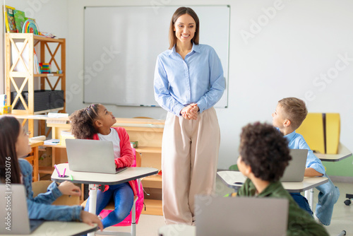 Fotografia Pupils listening their female teacher, sitting at desks with laptops, showcasing