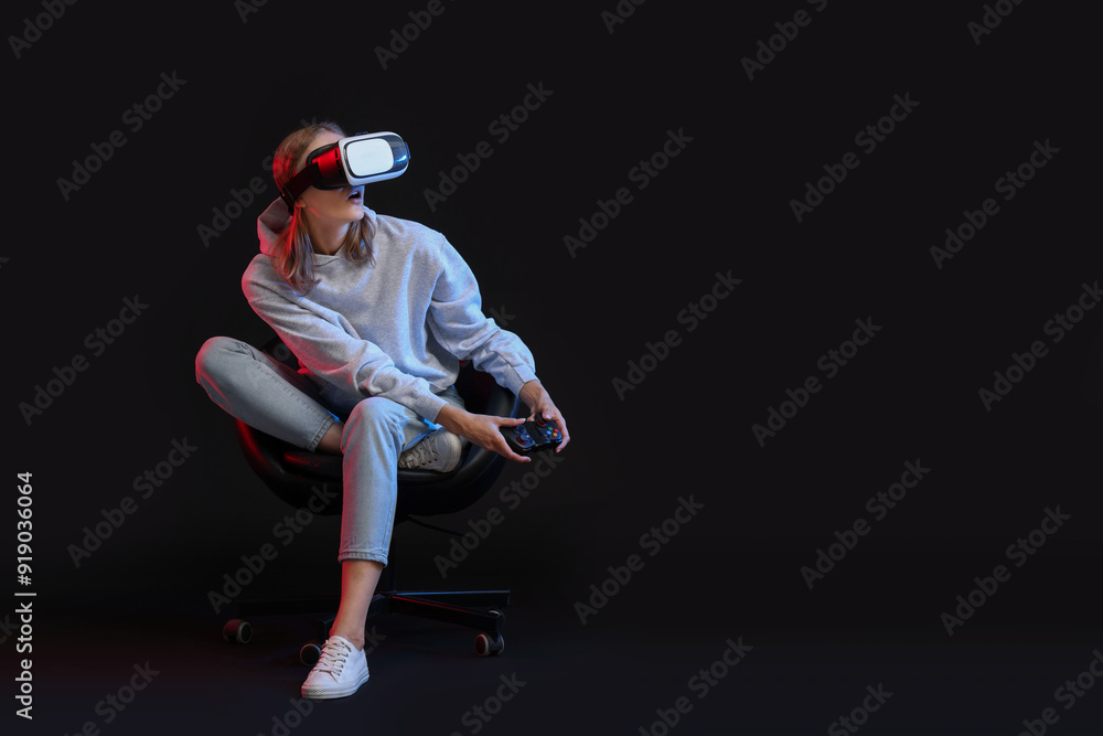 Emotional woman with virtual reality headset and controller sitting on chair in neon lights against black background, space for text Emotional woman with virtual reality headset and controller sitting on chair in neon lights against black background, space for text