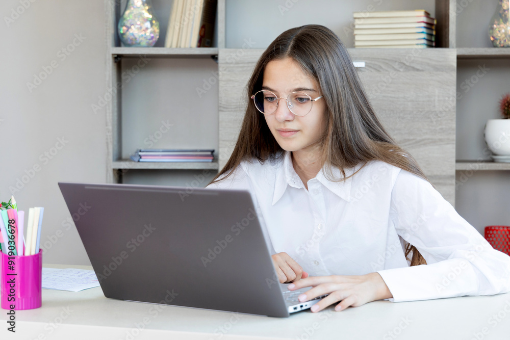 A teenage girl is studying using a laptop at home. A teenage girl with long brown hair and glasses is focused on her laptop while sitting at a desk in a home setting. A teenage girl is studying using a laptop at home. A teenage girl with long brown hair and glasses is focused on her laptop while sitting at a desk in a home setting.