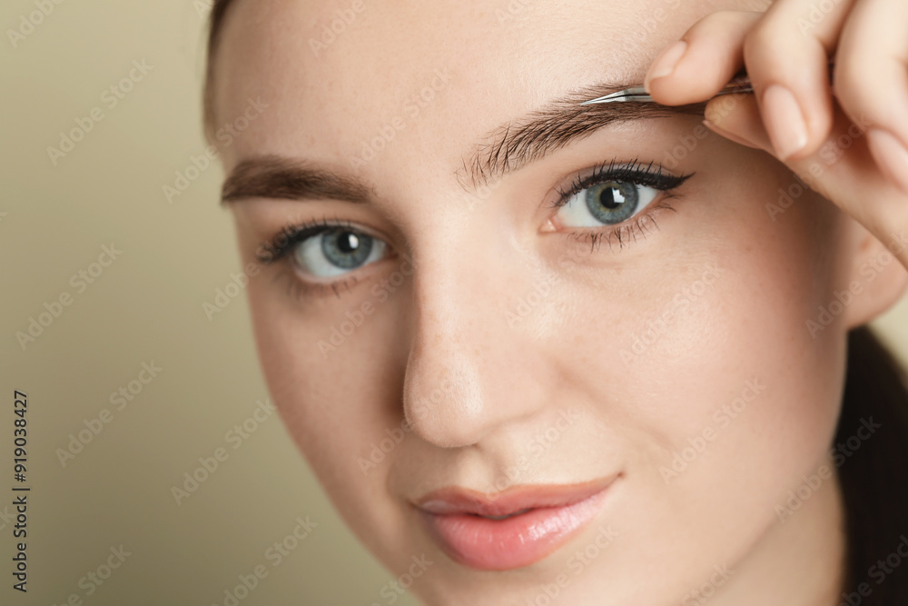 Fototapeta premium Young woman plucking eyebrow with tweezers on beige background, closeup
