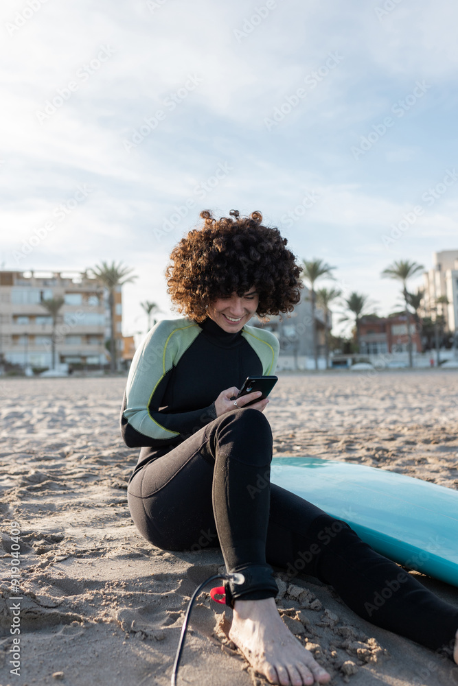 Fototapeta premium Smiling female surfer in wetsuit sitting on sandy beach