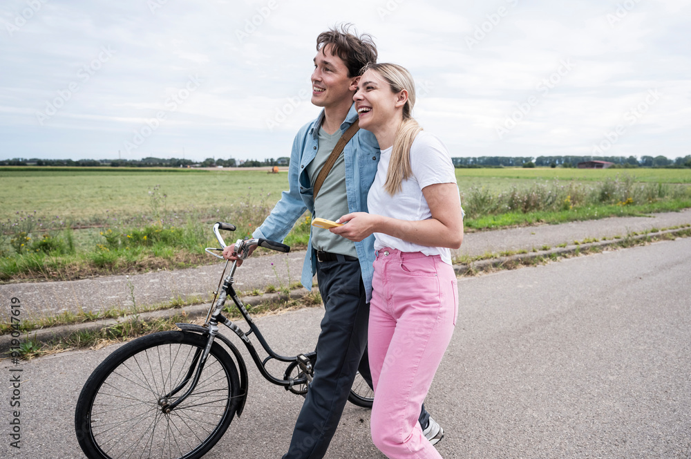 Happy young couple walking with bicycle on road near meadow Happy young couple walking with bicycle on road near meadow