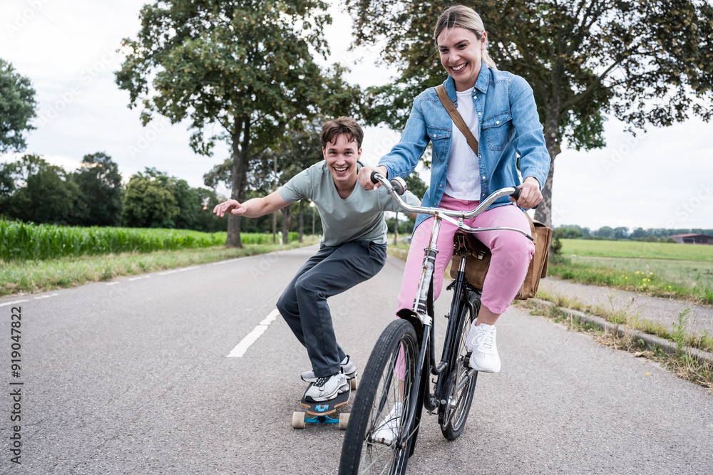 Happy woman riding bicycle with boyfriend skateboarding on road Happy woman riding bicycle with boyfriend skateboarding on road