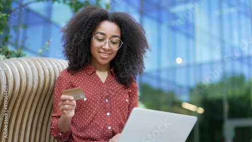 Satisfied black woman shopping online using laptop and credit card sitting on a bench near an office business building. Young African American female enjoying purchasing items conveniently outdoors photo