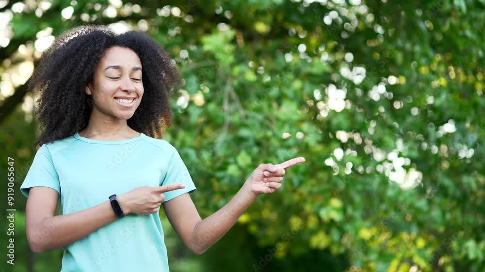 Portrait of smiling african american sportswoman pointing fingers at empty copy space for writing text and looking camera. Young sporty black woman standing in urban city park Template for advertising