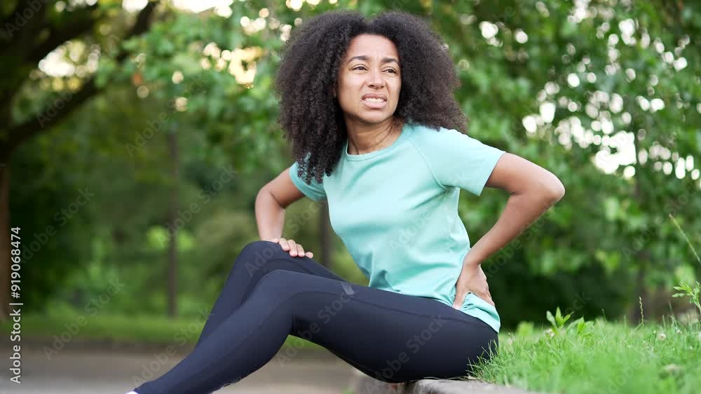 Young african american sportswoman suffering from back pain, sports injury, muscle spasm sitting in city park. Black woman having back flank ache, problem after exercise outside, massaging a sore spot Young african american sportswoman suffering from back pain, sports injury, muscle spasm sitting in city park. Black woman having back flank ache, problem after exercise outside, massaging a sore spot
