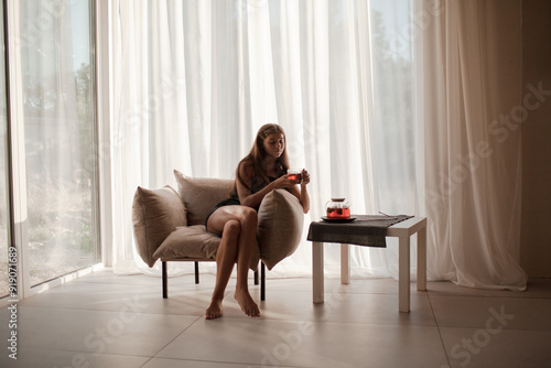 a young girl hugs her cat sitting in a chair at home. The female dressed s sweater. Cozy atmosphere at home with cup of tea