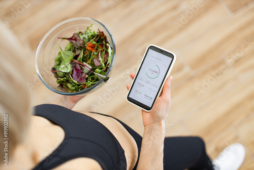 Quadro su tela Woman in fitness clothing tracking daily calorie intake on smartphone while holding bowl of fresh salad
