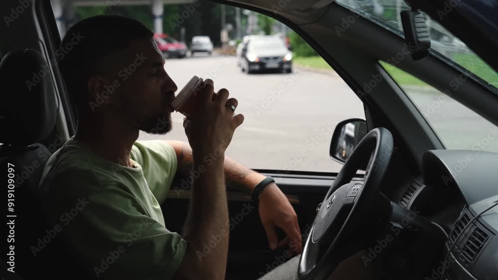 Male car traveler sits in a car and drinks coffee from a paper cup in the parking lot of a gas station. A young Caucasian man with a beard took coffee away, taking a break during a road trip Male car traveler sits in a car and drinks coffee from a paper cup in the parking lot of a gas station. A young Caucasian man with a beard took coffee away, taking a break during a road trip