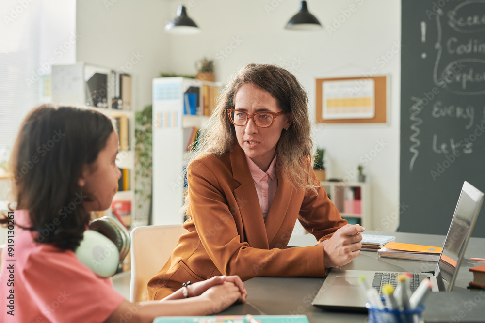 Fototapeta premium Portrait of adult woman as female teacher talking to girl during consultation in school classroom