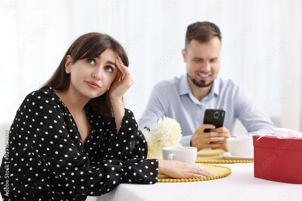 Embarrassing date. Bored woman sitting at table with man using smartphone indoors, selective focus Embarrassing date. Bored woman sitting at table with man using smartphone indoors, selective focus