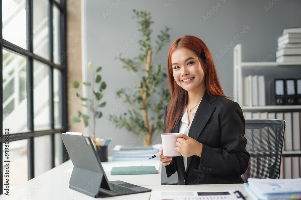 Asian businesswoman is smiling while holding a coffee mug and working at her desk in an office Asian businesswoman is smiling while holding a coffee mug and working at her desk in an office