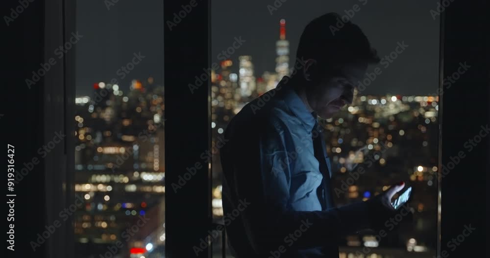 Businessman texting. Man standing near the windows at night urban background. Holding smartphone light reflecting on face. guy in suit standing by panoramic view of skyscrapers, Working later hours. Businessman texting. Man standing near the windows at night urban background. Holding smartphone light reflecting on face. guy in suit standing by panoramic view of skyscrapers, Working later hours.