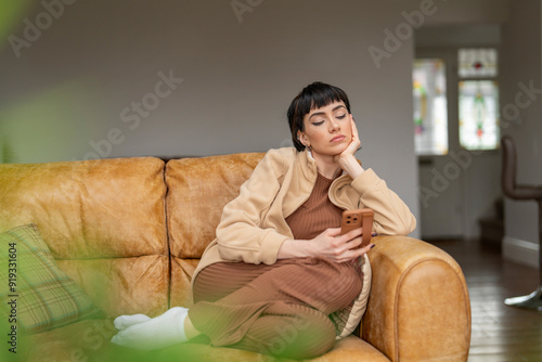 woman relaxes on cozy sofa, thoughtfully scrolling through her phone in warmly lit living room photo