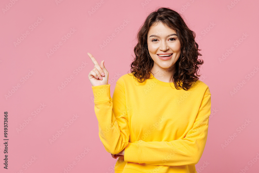 Young smiling cheerful happy woman she wearing yellow sweatshirt casual clothes pointing index finger aside on area mockup isolated on pastel light pink background studio portrait. Lifestyle concept. Young smiling cheerful happy woman she wearing yellow sweatshirt casual clothes pointing index finger aside on area mockup isolated on pastel light pink background studio portrait. Lifestyle concept.