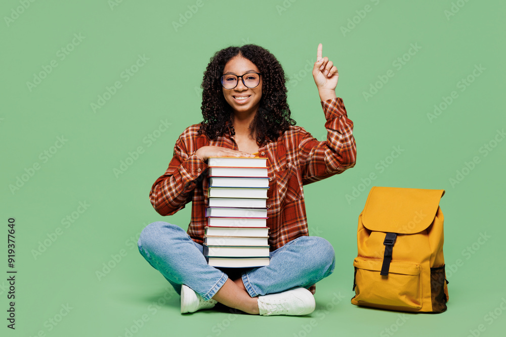 Full body young fun teen girl student wear brown yellow casual clothes backpack bag sits hold pile of books point finger up isolated on plain green background. High school university college concept.