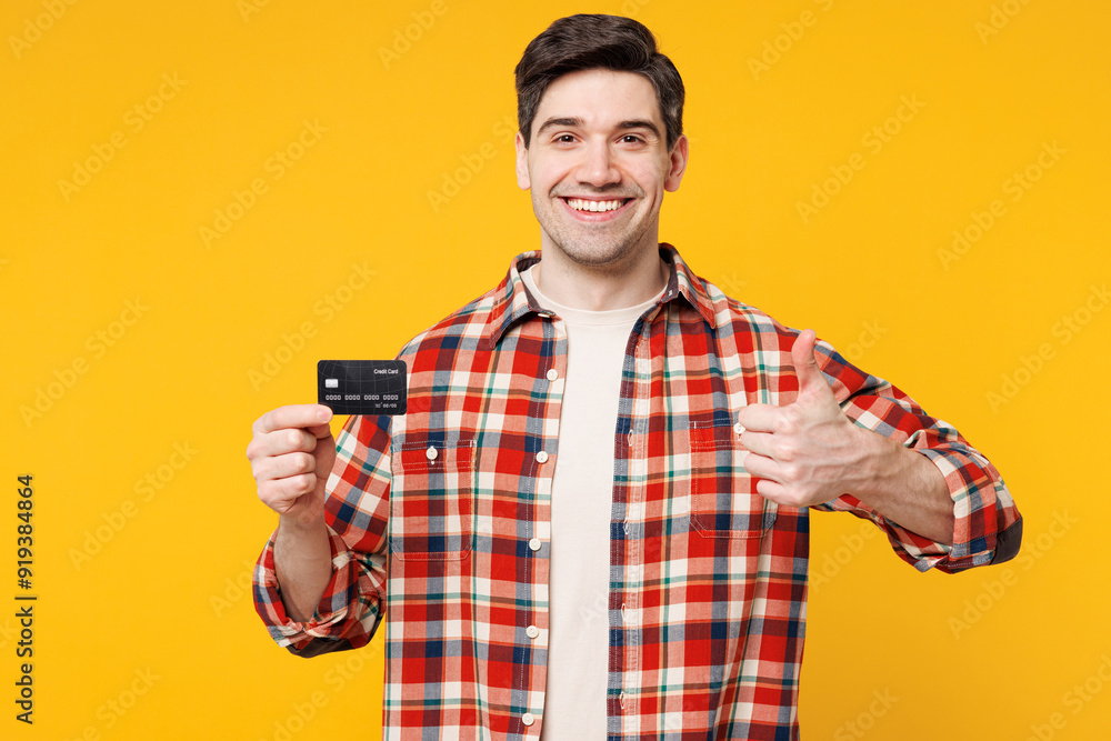 Young smiling happy man wears red checkered shirt casual clothes hold in hand mock up of credit bank card show thumb up isolated on plain yellow orange background studio portrait. Lifestyle concept. Young smiling happy man wears red checkered shirt casual clothes hold in hand mock up of credit bank card show thumb up isolated on plain yellow orange background studio portrait. Lifestyle concept.