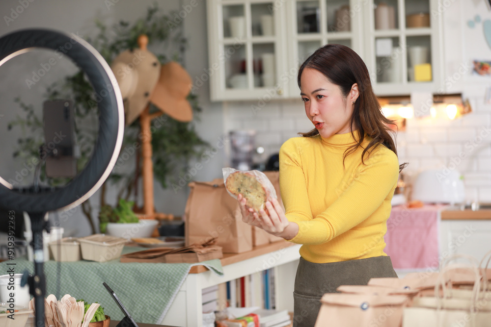 Female vlogger in bright yellow sweater enthusiastically presents food product in cozy kitchen. Captured mid-presentation through ring light, with smartphone positioned in front of her for recording Female vlogger in bright yellow sweater enthusiastically presents food product in cozy kitchen. Captured mid-presentation through ring light, with smartphone positioned in front of her for recording