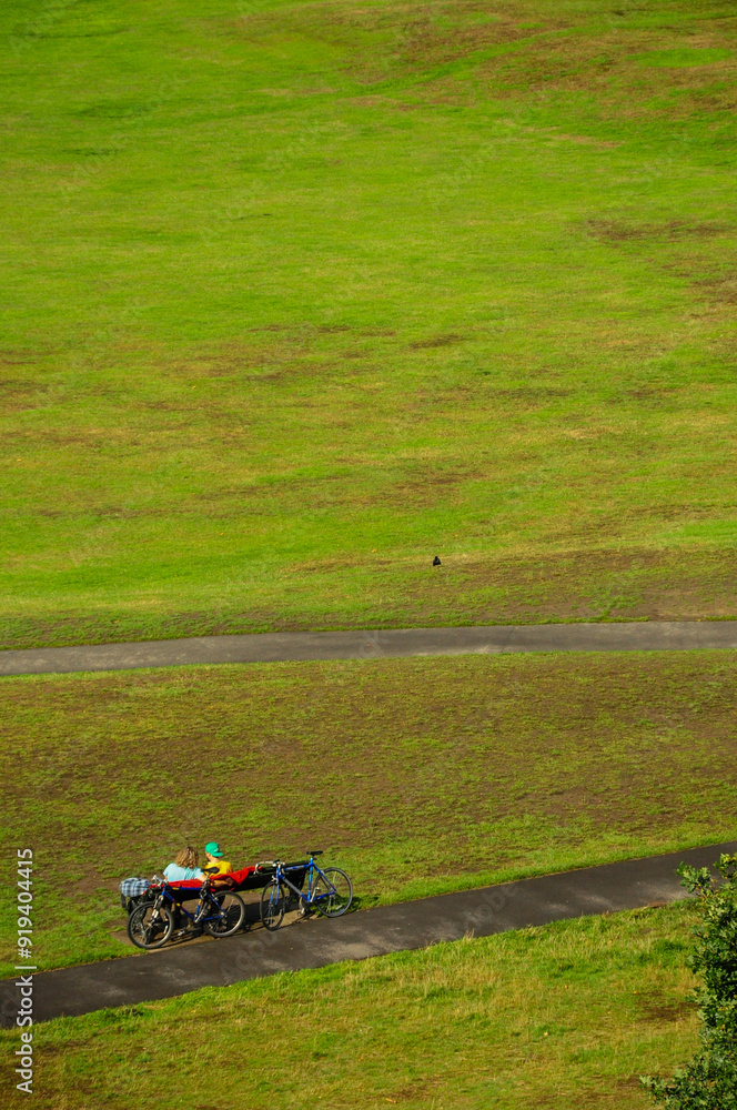 Aerial view of cyclists resting on a paved path in a lush, green parkland area. Aerial view of cyclists resting on a paved path in a lush, green parkland area.