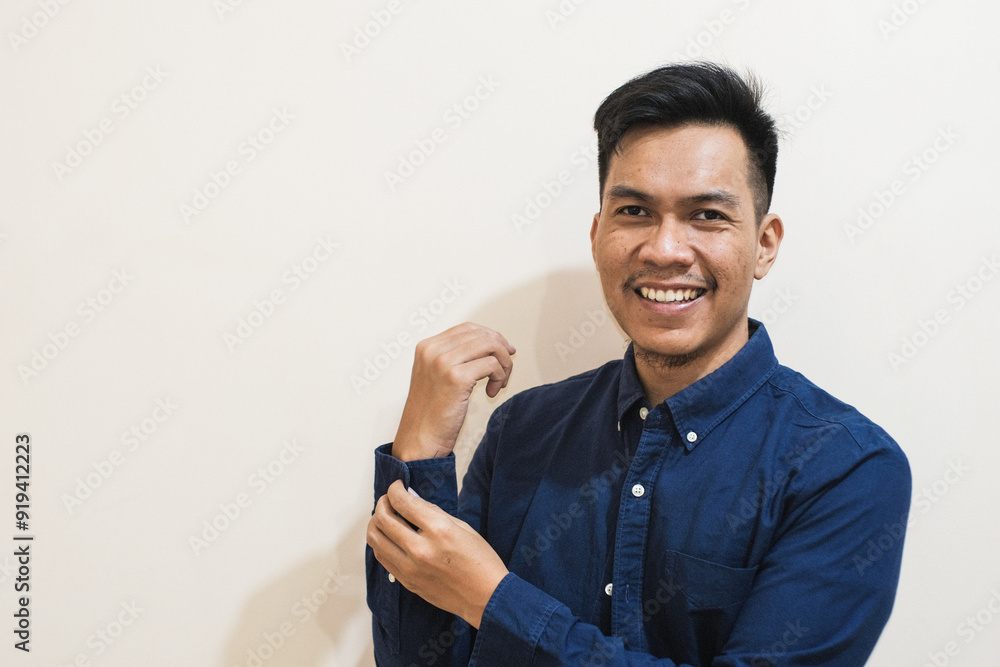 A Southeast Asian man smilling buttons up his shirt sleeves to get dressed, getting ready for work in the morning against a cream background. A Southeast Asian man smilling buttons up his shirt sleeves to get dressed, getting ready for work in the morning against a cream background.