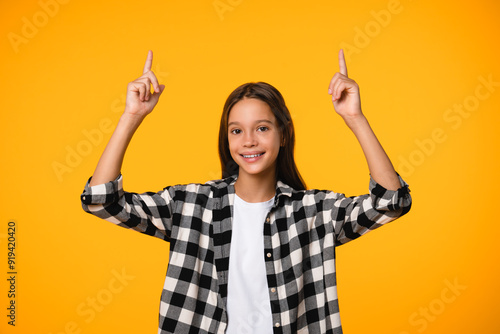 Smiling caucasian girl showing copy space pointing with her fingers isolated in yellow background. Schoolgirl teenager in plaid shirt showing advert place.