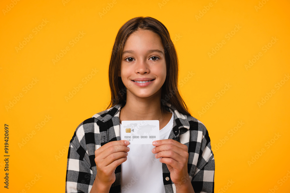Little smiling kid teenager child school girl 12-13 years old wearing white t-shirt hold credit bank card look at camera isolated on bright orange yellow background . Childhood online banking concept Little smiling kid teenager child school girl 12-13 years old wearing white t-shirt hold credit bank card look at camera isolated on bright orange yellow background . Childhood online banking concept