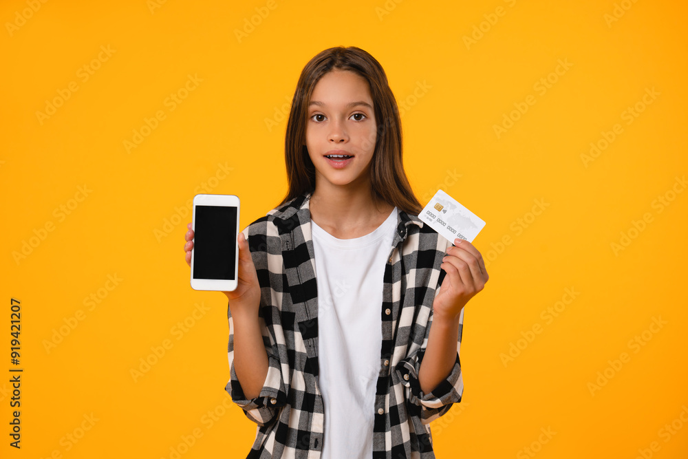 Shocked impressed surprised caucasian teenager girl schoolkid pupil student using smart phone with mockup blank screen paying with credit card isolated in yellow background. Shocked impressed surprised caucasian teenager girl schoolkid pupil student using smart phone with mockup blank screen paying with credit card isolated in yellow background.