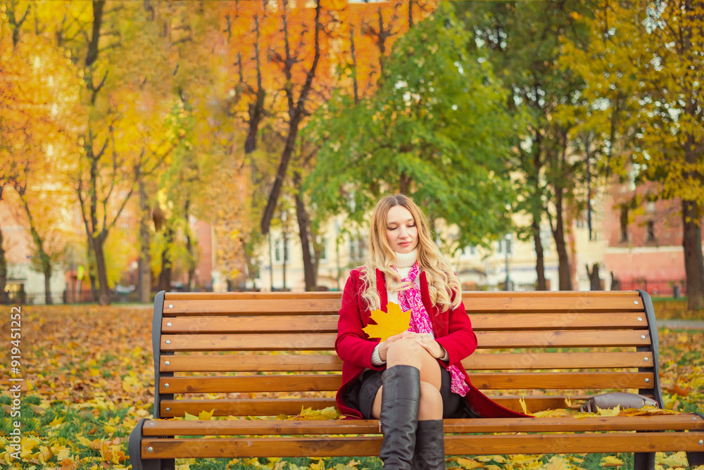 A girl in a red coat sits on a bench in an autumn park. She holds yellow leaves in her hand. A girl in a red coat sits on a bench in an autumn park. She holds yellow leaves in her hand.